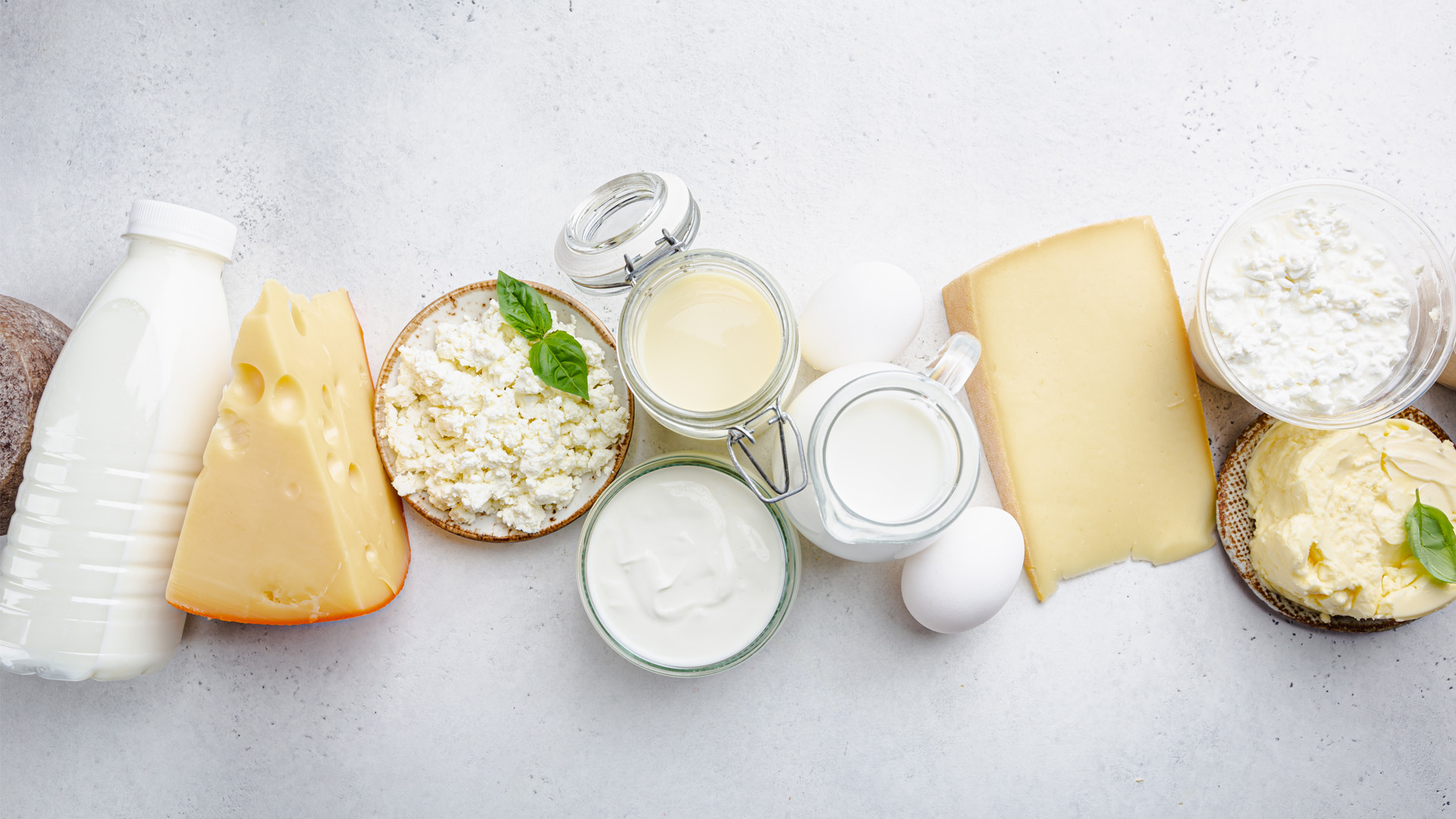 Varietà di prodotti lattiero-caseari su sfondo chiaro: latte, formaggi, yogurt ed uova.